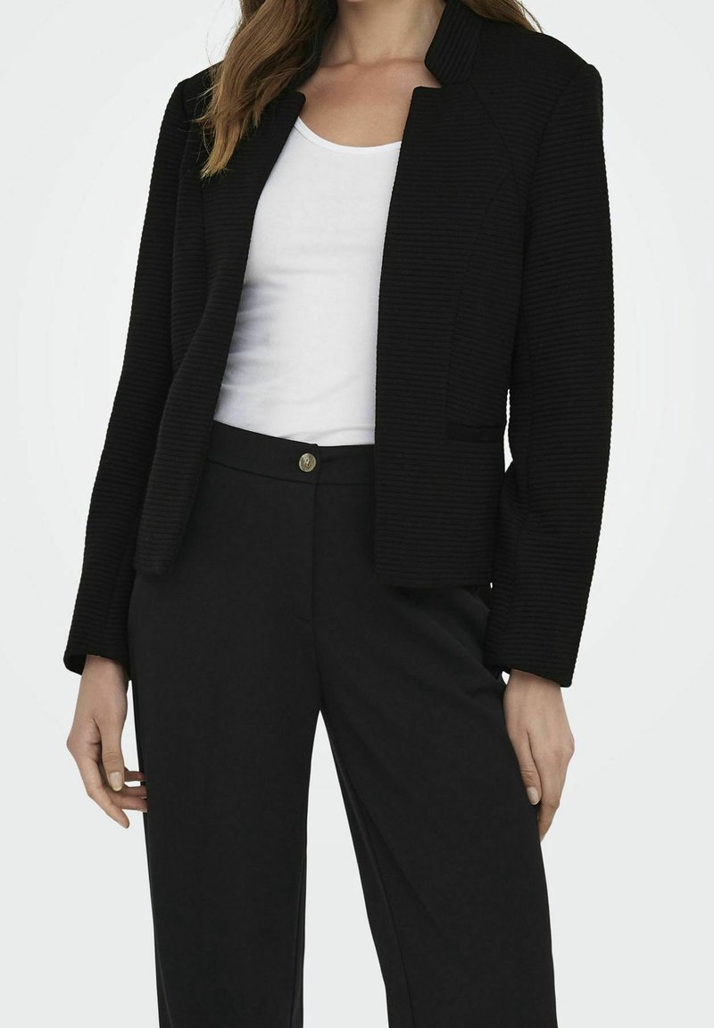 Femme portant un blazer noir texturé sur un top blanc, associé à un pantalon noir taille haute, devant un fond clair uni.