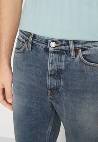 Jeans in denim blu scuro con un taglio classico, leggera sbiaditura, minuteria in bronzo e dettagli cuciti su tasche e cuciture.