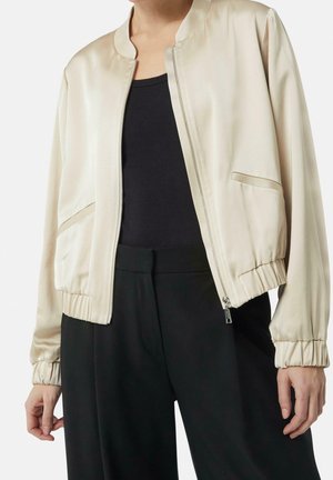 Beige satin bomber jacket with front zipper, side pockets, and elastic cuffs worn over a black top and black pants.