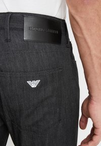 Černé džíny mají na pase koženou nášivku s nápisem "EMPORIO ARMANI" a bílé logo orla na zadní kapse.