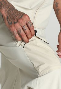 Pantaloni tecnici beige con tessuto testurizzato, tasca laterale con cerniera e dettagli neri a contrasto. Mano che afferra la tasca.
