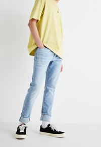 Ljusgul kortärmad skjorta som matchas med ljusblå slim-fit jeans, uppvikta ärmar, och svarta sneakers med vita sulor och snören.