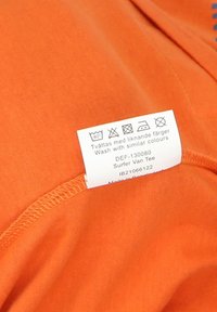 Oranssi puuvillapaita, jossa on hoito-ohjeita ja tuotetietoja näyttävä etiketti, joka on sileä ja pyöreä kaula-aukko.