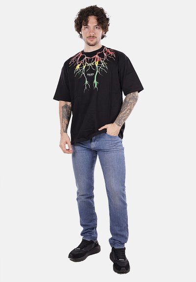 T-shirt nera oversize con grafica colorata a forma di radice sul petto, abbinata a jeans azzurri chiari e sneakers nere.