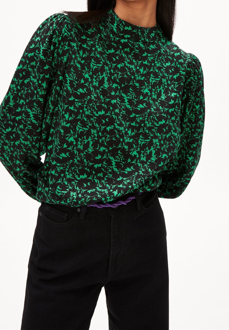 Femme portant un chemisier à manches longues à motif vert et noir, rentré dans un pantalon noir taille haute avec une ceinture tressée violette.