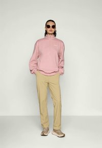 Ružový zipsový fleece pulóver s dlhými rukávmi, béžové nohavice a hnedé tenisky. Modelka nosí slnečné okuliare a stojí na neutrálnej pozadí.