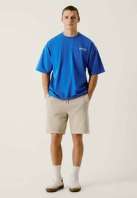 Zils kokvilnas T-krekls ar īsām piedurknēm un logotipu uz krūšu daļas, kombinēts ar beigu šortiem, baltiem zeķēm un baltām ikdienas kurpēm ar brūnām zolēm.