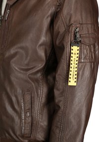 Brun læder bomberjakke med en lynlåslomme på ærmet og et gult mærke. Har en glat tekstur og en tætsiddende design.