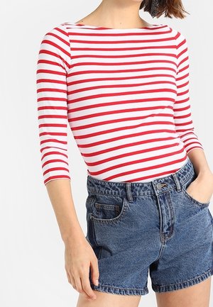 Kobieta w czerwono-białej paskowanej bluzce z długim rękawem i wysokim stanem niebieskich dżinsowych szortach, z rękami częściowo wsuniętymi w kieszenie.