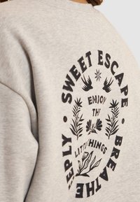 Sudadera gris con un gráfico circular negro que incluye el texto "SWEET ESCAPE" y diseños florales, que incluyen pájaros y hojas.