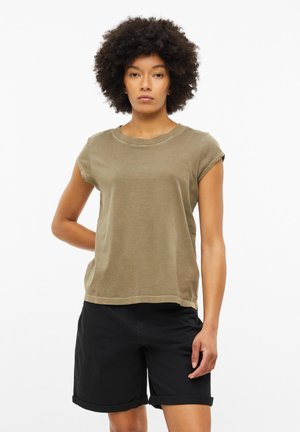 Młoda osoba z kręconymi włosami, ubrana w khaki t-shirt i czarne podwinięte szorty, stojąca z jedną ręką za plecami.