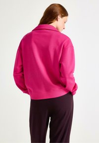 Kvinde med rødt hår iført en lys pink langærmet trøje og mørke bukser, stående med hænderne i lommerne, vendt væk fra kameraet mod en ensfarvet baggrund.