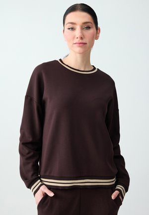 Femme portant un sweat-shirt marron foncé avec des poignets et un ourlet beige à rayures, les mains dans les poches, sur un fond clair uni.