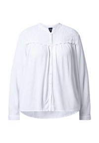 Camicia bianca con bottoni, dotata di una spallina strutturata, maniche lunghe e orlo arrotondato. Presenta un tessuto liscio con un aspetto leggermente stropicciato.