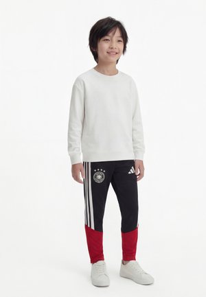 Smilende gutt som står, iført en hvit genser, svarte Adidas treningbukser med hvite striper og røde detaljer, og hvite joggesko på ensfarget bakgrunn.