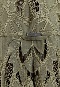 Olivgrünes Kleidungsstück mit aufwendiger Blattmuster-Stickerei, ausgeschnittenen Spitzenakzenten und einem silbernen Metalletikett mit der Aufschrift "ALLSAINTS."