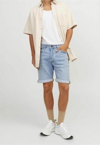 Man wearing beige short-sleeve shirt over white T-shirt, light blue rolled-up denim shorts, beige socks, and white sneakers standing against white background.