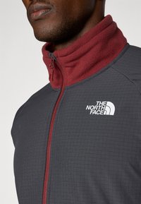 Grå fleecejakke med en maroon krave, med lynlåslukning og The North Face-logo i hvid. Struktureret gittermønster på overfladen.
