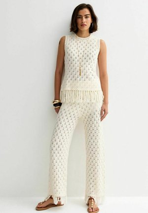 Kobieta ubrana w kremowy, bezrękawnik z szydełkowym wzorem z frędzlami i dopasowane szerokie spodnie, uzupełnione bransoletkami, naszyjnikiem i sandałami.