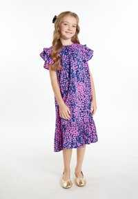 Rožinės ir mėlynos leopardų rašto suknelė su raukšlėtomis rankovėmis ir kelių lygių apatiniu kraštu. Modelis vilki auksines slidžias batus, stovintis ant balto fono.