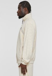 Kauris fleece vaatetakk, millel on kõrge krae, elastsed mansetid, küljetaskud ja peene reljeefne muster. Rinnal on väike logo.