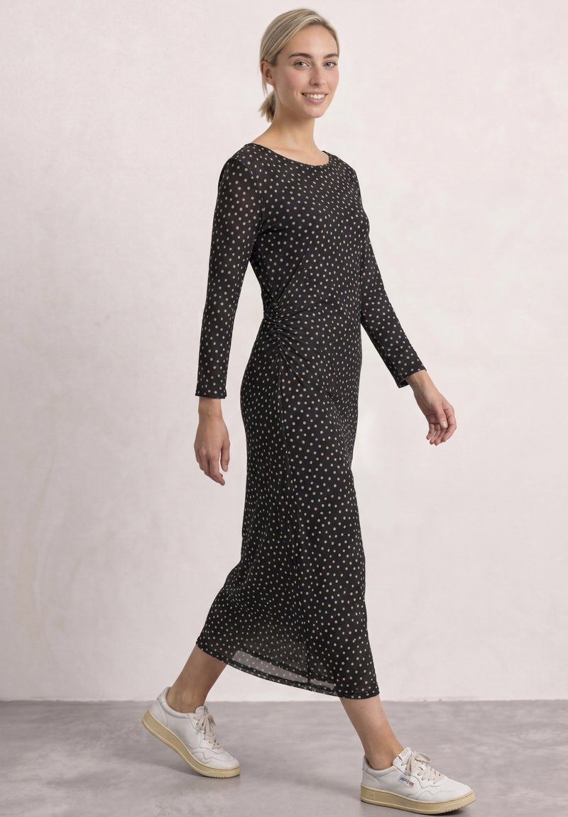Femme marchant dans une robe midi noire à manches longues avec des pois blancs, portant des baskets blanches, souriante, les cheveux attachés en arrière.