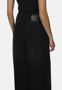 Zwarte wijde jeans met hoge taille, twee achterzakken en een leren logo patch. De stof heeft een gladde textuur en een donkere afwerking.