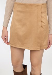 Minigonna in finta pelle scamosciata color tan con design a incrocio, linee pulite e cuciture sull'orlo. Abbinata a un top a maniche lunghe a coste e stivali marroni al ginocchio.