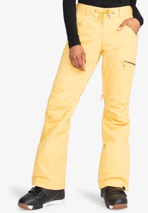 Gele sneeuwbroeken met een ruime pasvorm, voorzien van ritszakken, een trekkoord in de taille en versterkte zoom. Gepaard met zwarte snowboardlaarzen.
