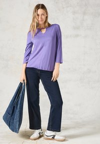 Top viola a maniche lunghe con dettaglio a chiave, abbinato a jeans a gamba larga blu scuro. Il modello tiene una borsa in denim e indossa sneakers bianche.