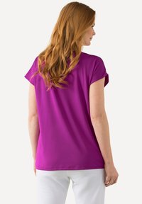 Krátký rukáv, fialový top vyrobený z hladké látky, s volným střihem a jemným detailem výstřihu. Zadní strana je jednoduchá s zakřiveným lemem.