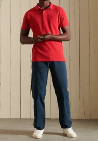 Polo shirt rouge à manches courtes et col, associé à un pantalon bleu marine et des baskets blanches. Design simple sans motifs distincts.