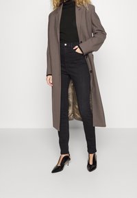 Long brown coat over a black turtleneck, paired with high-waisted black jeans and black heeled shoes. Textured lining visible inside the coat.