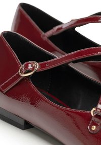 Ballerine in vernice bordeaux con design a T, caratterizzate da una fibbia in tono dorato e un tacco nero basso. L'interno ha una texture liscia.
