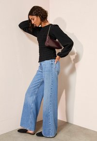 Czarna bluzka z długimi rękawami, zestawiona z jasnoniebieskimi dżinsami o szerokich nogawkach i czarnymi butami wsuwanymi. Trzyma bordową torebkę na łańcuszku.