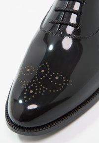 Chaussure en cuir verni noir à bout pointu, présentant des motifs décoratifs perforés et des lacets noirs. Texture lisse avec finition brillante.