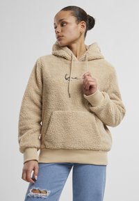 Žena v béžovej huňatej mikine s predným vreckom a svetlomodrých roztrhaných džínsach, pozerá sa doľava na jednoduchom pozadí.