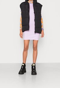 Černá péřová vesta se zipem, nošená přes světlou fialovou upnutou šaty. Černé robustní boty s výraznými akcenty doplňují celý outfit.