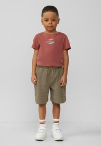Rode katoenen T-shirt met grafisch ontwerp, gecombineerd met lichtbruine shorts met zakken, en witte sneakers met rode gestreepte sokken.