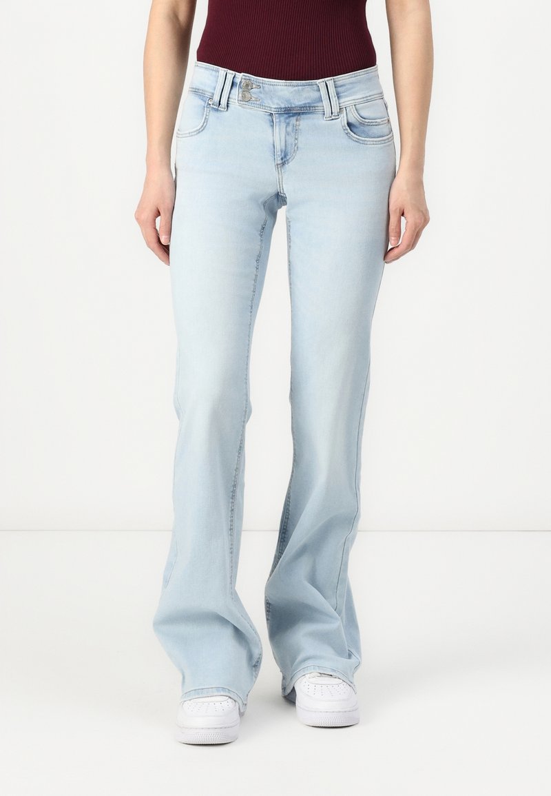 Jean flare bleu clair en denim doux, présentant un design à cinq poches et des accents en métal argenté. Porté avec un haut bordeaux et des baskets blanches.