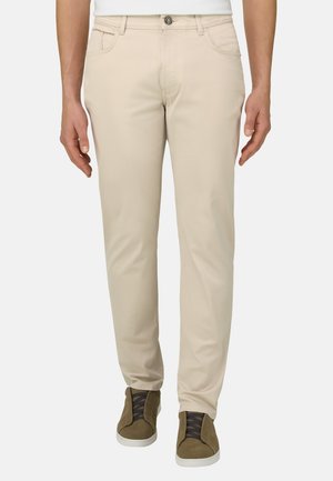 Mann trägt beige, eng anliegende Hose und olivgrüne Sneaker mit schwarzen Schnürsenkeln, steht vor einem einfarbigen Hintergrund.