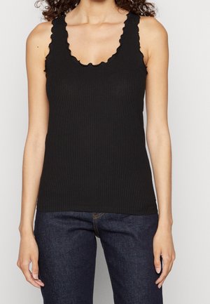 Vrouw draagt een zwarte geribbelde mouwloze top met gegolfde randen en donkerblauwe spijkerbroek, staand tegen een effen lichte achtergrond.