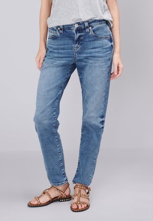 Onderlichaam van een persoon die lichtblauwe vervaagde spijkerbroek, een grijs topje en bruine platte sandalen met studs draagt tegen een effen achtergrond.