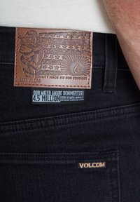 Jeans de denim negro con un parche de cuero marrón texturizado que presenta un diseño en relieve y una etiqueta ecológica que destaca el ahorro de agua.