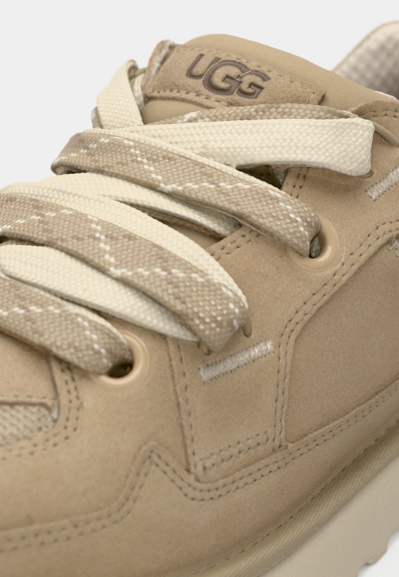 Beige Schnürsenkel in tanfarbenen Wildleder-Sneakern, mit einer strukturierten Oberfläche und gestickten Akzenten. UGG-Logo auf der Zunge angebracht.