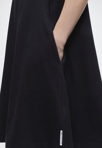 Δεξί χέρι που βγαίνει στην πλευρική τσέπη ενός μαύρου φορέματος μήκους μέχρι το γόνατο με μια μικρή λευκή ετικέτα μάρκας κοντά στο τελείωμα.