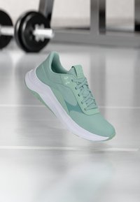 Αθλητικό παπούτσι ανοιχτού πράσινου χρώματος με λευκή σόλα, αιωρούμενο πάνω από το πάτωμα γυμναστηρίου, με θολές βάρη στο παρασκήνιο.