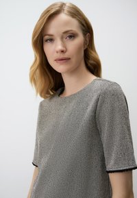 Blouse en chevrons gris texturé avec un col rond, des manches courtes et des finitions contrastantes aux bords des manches. Tissu au fini lisse.