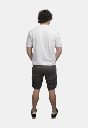 T-shirt bianco in cotone, maniche corte, abbinato a pantaloni cargo scuri con tasche laterali. Sneakers nere con dettagli verdi e arancioni.