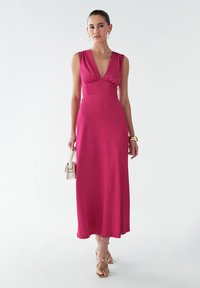 Magenta maxi dress with a deep V-neck, ruched shoulders, and a fitted waist. Worn with gold sandals and a small white handbag.